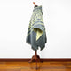 A green-and-cream patterned poncho drapes over a wooden stand, displayed against a plain white wall with a hardwood floor.