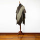 A mannequin displays a patterned poncho with green and brown geometric designs, draped in a standing position. It is set on a wooden stand in a room with hardwood flooring.