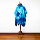 A blue tie-dye hooded poncho is displayed on a wooden mannequin stand, in a minimalist room with a wooden floor and white wall background.