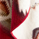 A woolen blanket with a red edge and geometric patterns drapes softly. The design includes shades of brown, red, and cream, showcasing a zigzag motif.