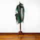 A green ombre shawl with intricate patterns drapes over a wooden mannequin. It stands on a polished wood floor against a plain white wall, creating a minimalist display.