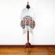 A white poncho with geometric patterns displayed on a stand, against a plain white wall and wooden floor.