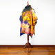 A colorful, tie-dye poncho is displayed on a wooden mannequin stand, standing in a room with a hardwood floor and a plain white wall.