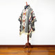 A patterned poncho, displayed on a mannequin, features geometric designs in black, red, and beige. The poncho hangs in a room with a wooden floor and a plain white wall.
