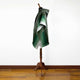 A green cloak with gradient tones drapes over a dark wooden mannequin stand, set against a white wall and wooden floor in a minimalistic indoor space.