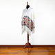 Poncho displayed on a wooden stand, featuring white fabric with decorative geometric and floral patterns in red and black. Set against a plain, white wall and wooden floor.
