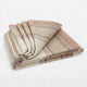 Zhumir baby alpaca wool blanket – a folded blanket rests neatly, featuring a pattern of parallel stripes in shades of beige, rust, and gray. The background is a plain, neutral surface
