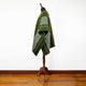 A green poncho with white and brown stripes is draped over a mannequin stand, set against a plain white wall and wooden floor.