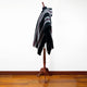 A mannequin displays a black, gray, and red-striped hooded poncho, standing on a wooden floor against a plain white background.