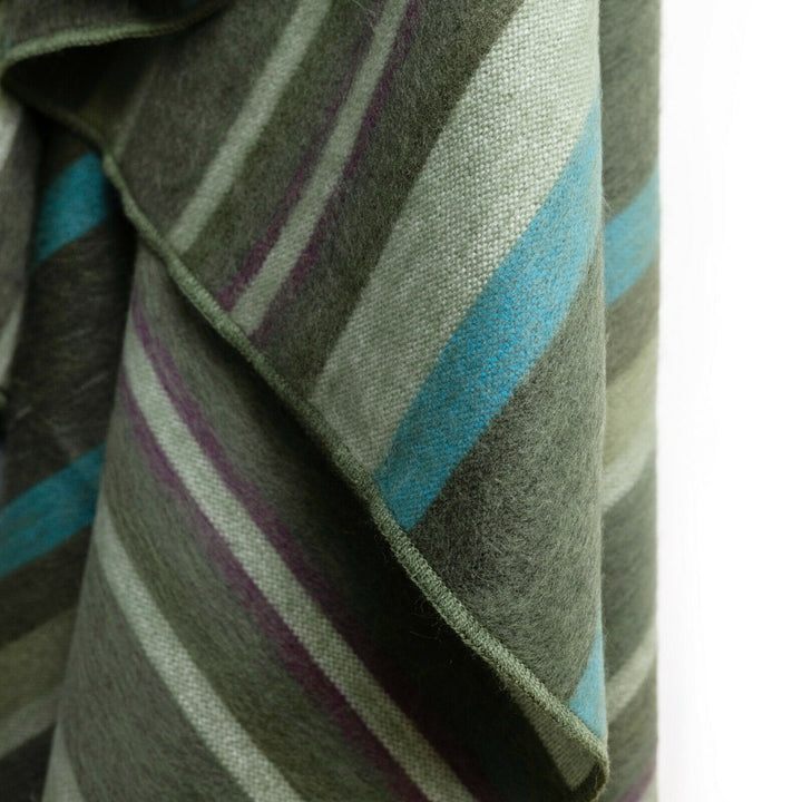 Jambue baby alpaca wool blanket – a woolen fabric with green, blue, and purple stripes drapes downward, showcasing its soft texture. The close-up view highlights the fine weaving detail, with no background distractions