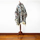 A patterned poncho with geometric designs hangs on a wooden mannequin stand in a room with a hardwood floor and a plain white wall.