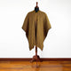A striped poncho with yellow, brown, and black patterns is displayed on a mannequin. It drapes elegantly over a wooden floor against a plain white wall.