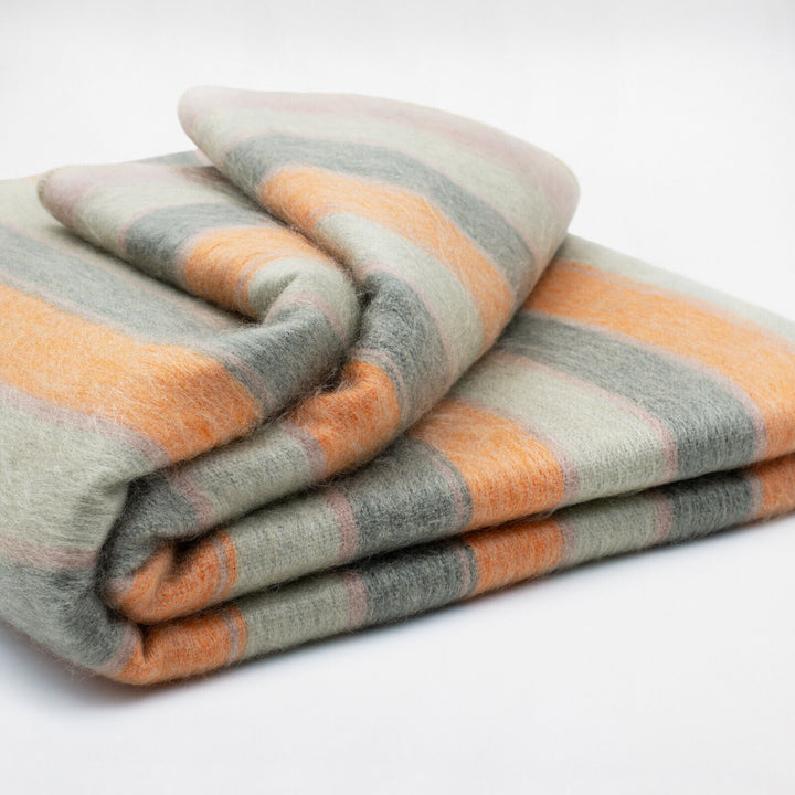Tumbunuma baby alpaca wool blanket – a folded, multicolored woolen blanket with broad orange, gray, and green stripes rests on a plain white surface