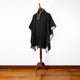A black poncho with embroidered collar detailing is draped over a mannequin, standing on polished wooden flooring against a plain white wall.