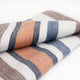 Sopoto baby alpaca wool blanket – a folded, striped woolen blanket with shades of orange, blue, and gray, lies on a plain white surface, showcasing its soft texture and woven pattern