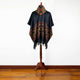 A poncho with geometric patterns in red and yellow hangs on a wooden stand. The poncho's fringe drapes near the floor in a room with a wooden floor and white walls.