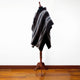 A brown poncho with white and gray diagonal stripes drapes over a mannequin. The setting features a light-colored wall and wooden floor, creating a minimalist display.