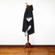 A dark cloak with a hood drapes over a wooden mannequin, standing on a polished hardwood floor against a plain white wall.