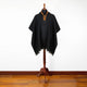 A dark poncho with ornate embroidery displayed on a wooden mannequin stand, situated on a polished wooden floor against a plain white backdrop.