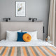 Tumbunuma baby alpaca wool blanket – bed with white pillows, blue and orange cushions, and orange-striped blanket beneath a framed bird illustration. Two black wall-mounted lamps and a small side table with a lantern are present