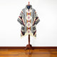 Poncho with geometric patterns, displayed on a wooden stand. The design features red, black, and cream colors, with fringed ends. Set against a plain white wall and hardwood floor.