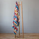 A colorful, patterned blanket hangs on a simple wooden stand. The scene is set against a light gray wall in a room with parquet flooring, creating a minimalistic atmosphere.