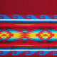 A brightly colored woven textile features geometric patterns in red, blue, yellow, and turquoise, showcasing intricate designs and zigzag motifs against a predominantly red background.