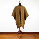 A mannequin displays a striped, multicolored poncho, featuring yellow and brown diagonal lines. It stands in a room with white walls and wooden flooring, conveying a warm, natural setting.