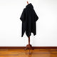 Achema llama wool hooded poncho – a black hooded cloak is displayed on a wooden mannequin stand, placed on a polished hardwood floor against a plain white background