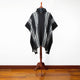 A black and gray-striped poncho hangs on a wooden stand. It features a hood and button, set against a minimalistic white wall and wooden floor background.