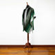 A green and gray poncho with intricate patterns is displayed on a wooden mannequin. It stands on a hardwood floor against a plain white wall.