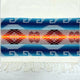 A woven textile features intricate geometric patterns in blue, orange, and brown hues, with diamond shapes and Greek key motifs. It is bordered by white fringe, lying on a flat surface.