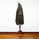 A dark brown tweed poncho drapes over a wooden dress form stand, set against a minimalistic white wall and polished wooden floor.
