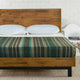 Jambue baby alpaca wool blanket – bed with striped blanket sits in a room with white brick walls. Two white pillows rest against the wooden headboard. A white chair holds books labeled DESSERT and another unreadable title