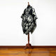 A black poncho with white geometric and dreamcatcher designs is displayed on a wooden stand. It rests on a wooden floor against a plain white wall.