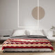A bed features a red and beige patterned blanket, resting neatly with white pillows. It's placed against a minimalist light gray wall, decorated with abstract lighting and a circular design.