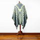 A woven poncho with intricate geometric patterns hangs on a wooden mannequin stand, set against a plain white background on a polished wooden floor.