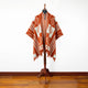 A colorful poncho with orange and white geometric patterns hangs on a mannequin, standing on a wooden floor against a plain white wall.