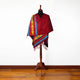 A mannequin is draped with a colorful shawl featuring geometric patterns. The shawl displays rich reds, blues, and yellows. It stands on a wooden floor against a plain white background.