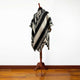 A black and grey poncho with geometric patterns hangs on a wooden mannequin stand, set against a white wall and wooden floor background.