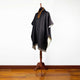 A dark poncho with a decorative collar is displayed on a wooden stand, set against a plain white wall and a wooden floor.