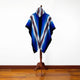 Wuachapa llama wool hooded poncho – a multicolored poncho with blue, white, and brown stripes hangs on a wooden stand against a white wall in a room with a wooden floor