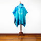 A blue tie-dye poncho with a hood hangs on a wooden mannequin stand, placed on a polished wooden floor against a plain white wall.