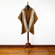 A brown poncho with white geometric patterns is displayed on a wooden mannequin stand against a white wall, on a polished wooden floor.
