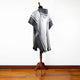 A gray and white patterned poncho with a hood drapes over a wooden mannequin stand, set against a plain white wall and wooden floor background.