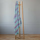 Tzacantza baby alpaca wool blanket – a wooden blanket rack stands upright, holding a colorful striped blanket draped over it, in a room with light walls and a parquet floor