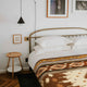 A neatly made bed with a geometric-patterned blanket and stacked white pillows is next to a small wooden side table. A pendant light hangs overhead; framed artwork decorates the white wall.