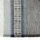 A woven textile features a central geometric pattern with diamond shapes, flanked by subtle blue and gray stripes. The context is a soft, fabric surface with fringed edges.