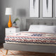 A bed with a patterned blanket lies in a modern bedroom. A gray tufted headboard complements a bedside table with an orange lamp and potted plant. Feather artwork adorns the wall.