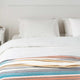 Nongora baby alpaca wool blanket – a neatly made bed with white pillows and blankets topped by a colorful striped throw. It's placed in a minimalistic white bedroom with a small bedside table